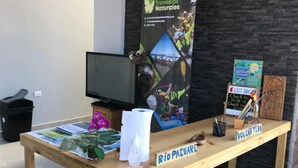 A wooden table with a sign reading "RIO PACUARE," a poster for "Travesias Naturales," a television, and a trash bin. - Hotel Pacuare Turrialba (Santa Rosa)