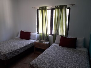 A room with two single beds, each with patterned bedding and red pillows. There is a small wooden nightstand with a lamp between the beds. A window with green curtains allows natural light into the room. - Familia Sosa Suazo (San José del Cabo)