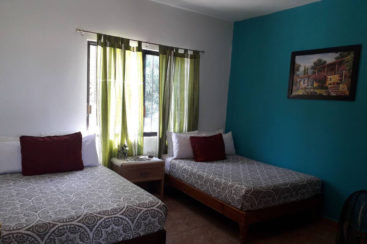 Una habitación con dos camas, una mesita de noche, una ventana con cortinas y un cuadro en la pared.