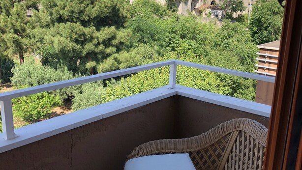 Un balcón con una silla de mimbre y un cojín blanco, con vistas a un pueblo en una ladera muy poblada con edificios de tejados de terracota.