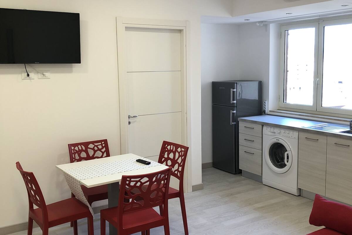 A compact living space with a dining area, a sofa, a washing machine, and a TV mounted on the wall.