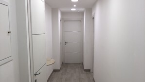 Un pasillo estrecho con paredes blancas y suelo de madera. Hay puertas a ambos lados, y un estante pequeño está fijado a la pared.
