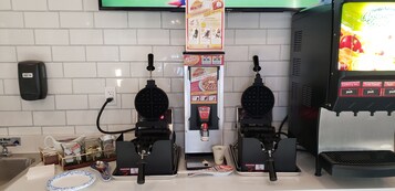 Una estación de comida autoservicio con máquinas para gofres, dispensador de bebidas y señal de precaución.