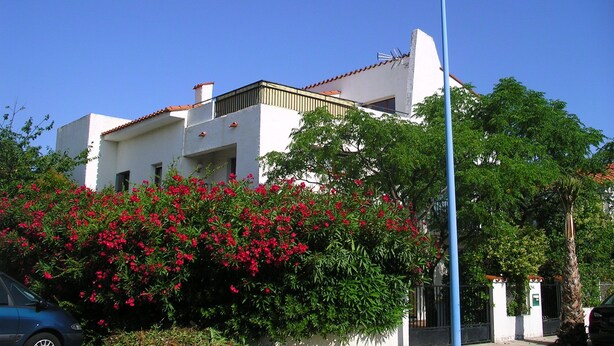 A white building with a red-tiled roof, surrounded by lush greenery and red flowers. - Large T3 loggia in house 500m from the beach (saint cyprien)