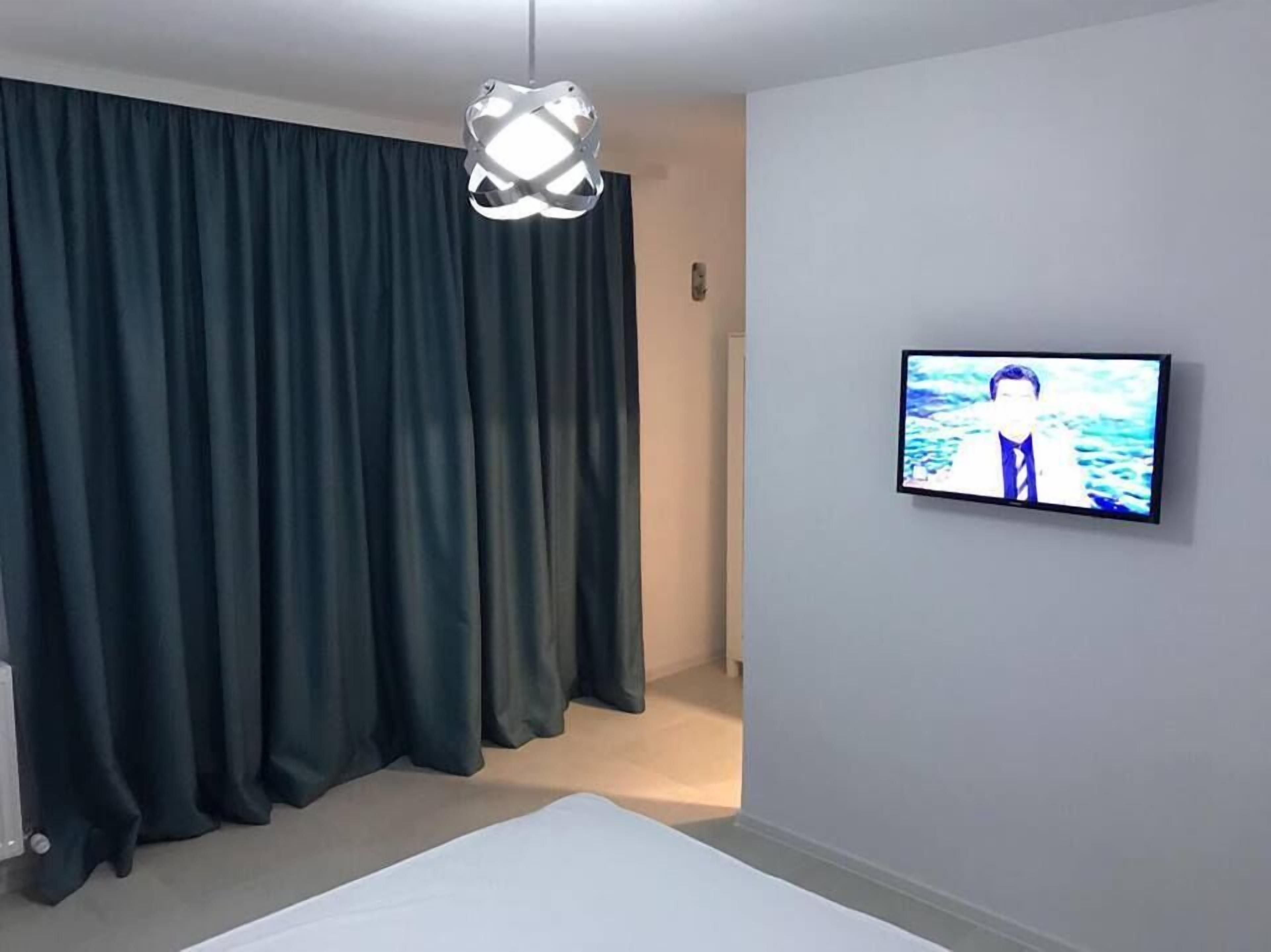 A modern hotel room with a large window, dark curtains, a flat-screen TV on the wall, and a unique pendant light.