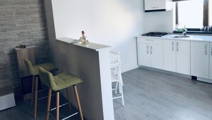 Una cocina moderna con alacenas blancas, estufa a gas y ventana con vistas. Hay dos taburetes verdes y una mesita con una botella.