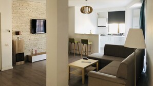 Una sala de estar moderna con un sofá, una mesa de centro y un televisor montado en una pared de ladrillo. Se ve un área de cocina con estufa y armarios al fondo.