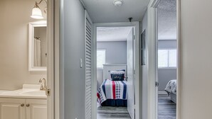 Ein Flur, der in ein Schlafzimmer mit einem Bett, einem Nachttisch und einer Nachttischlampe führt. Ein Fenster mit Jalousien, eine Tür und eine Leuchte über dem Spiegel sind ebenfalls vorhanden.