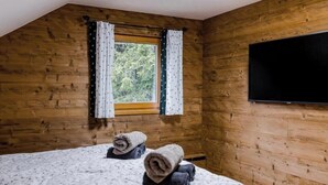 Ein Zimmer mit Holzwänden, einem großen Fenster mit Vorhängen, einem Bett mit aufgerollten Handtüchern und einem an der Wand montierten Flachbildfernseher.