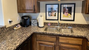 A kitchen with wooden cabinets, a granite countertop, a Keurig coffee maker, a coffee and friends sign, a paper towel holder, and a framed picture.