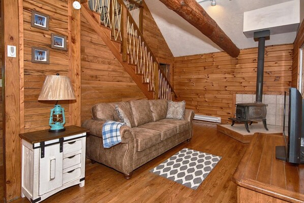 A cozy cabin interior with a wooden staircase, a sofa, a small cabinet, a heater, and a television. - Wildflower Cabin in Hocking Hills | Private, Cozy, Hot Tub Getaway (Rockbridge)