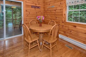 A wooden cabin interior with a round dining table surrounded by chairs, a vase with flowers, and a view of the forest through a glass door. - Wildflower Cabin in Hocking Hills | Private, Cozy, Hot Tub Getaway (Rockbridge)