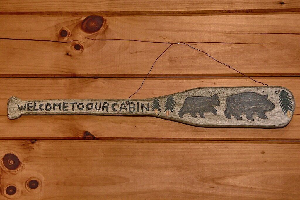 Ein hölzernes Schild mit Baseballschläger-Motiv, zeigt zwei Bären und den Schriftzug "WELCOME TO OUR CABIN".