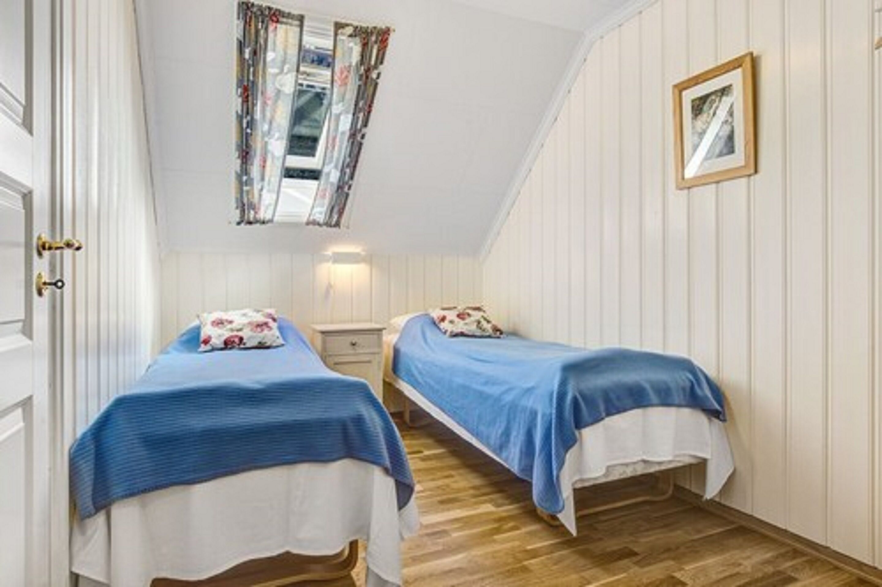 A bedroom with two single beds, a window with curtains, a bedside table, and a framed picture on the wall.