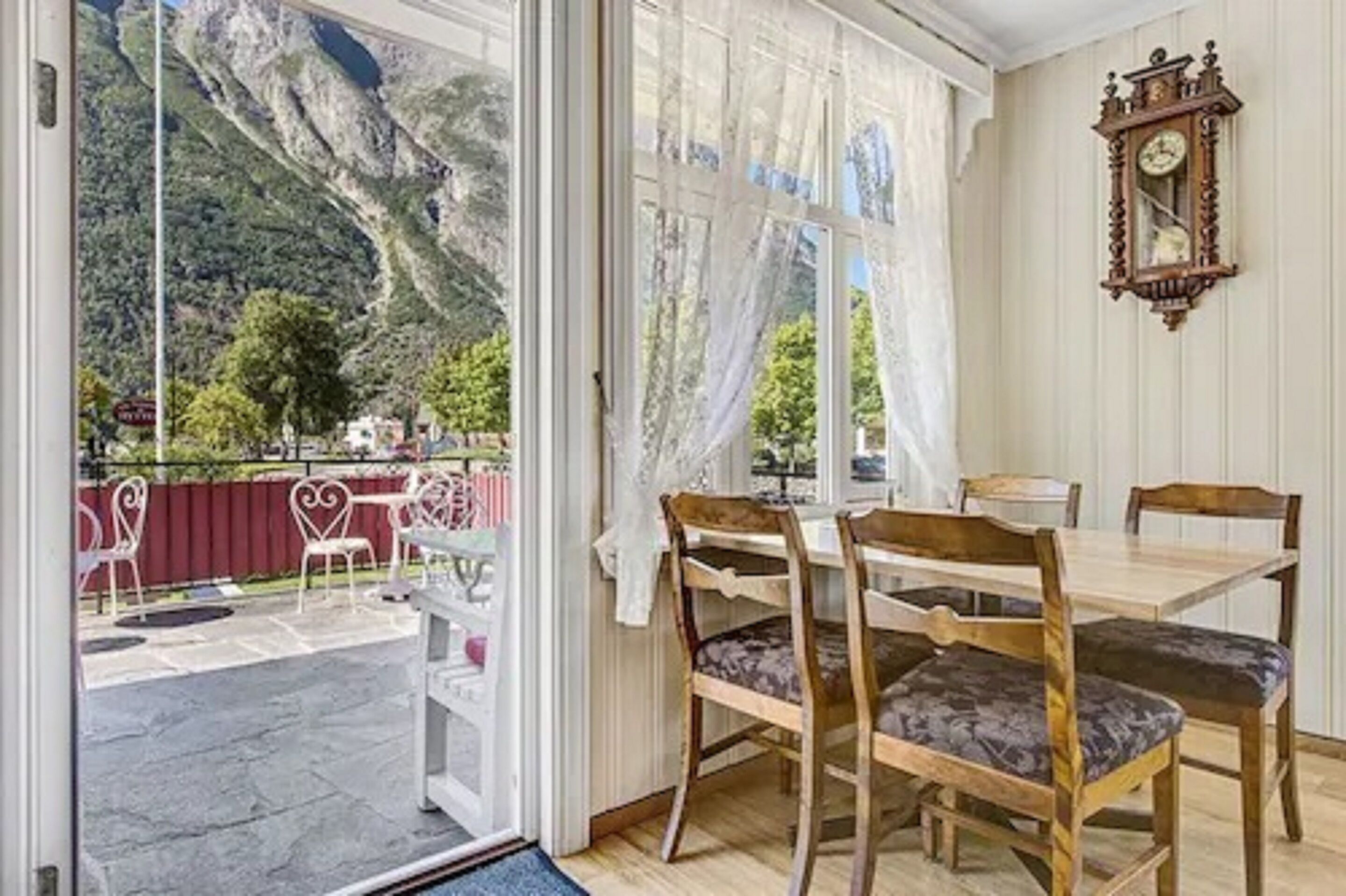Un coin repas avec une table et des chaises, une vue sur un paysage montagneux par une porte vitrée et une horloge fixée au mur.