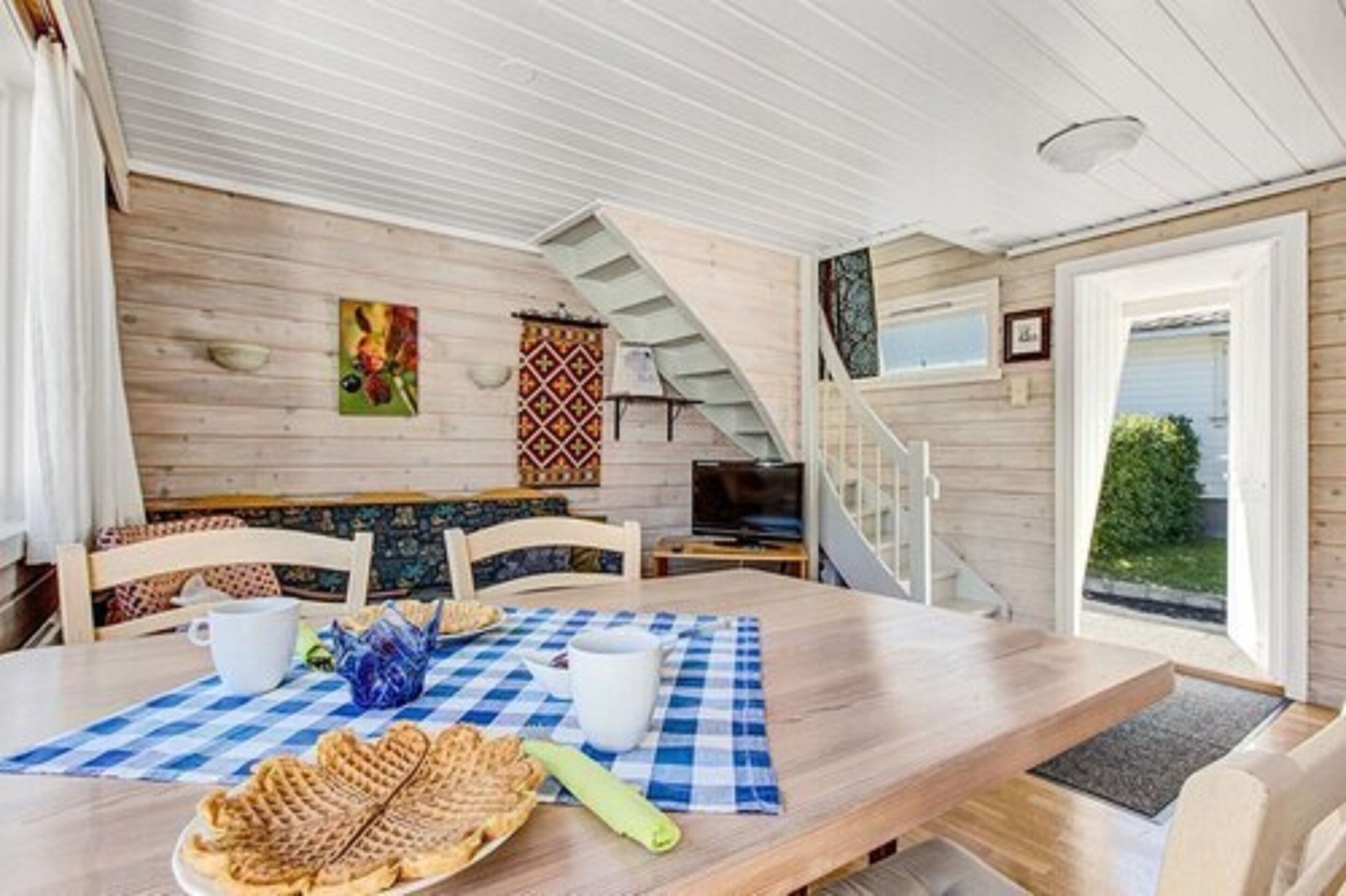 Interior acolhedor de uma cabana com uma mesa de jantar de madeira, cadeiras e toalha quadriculada. Uma escada leva a um andar superior, e uma janela oferece vista para o verde lá fora.