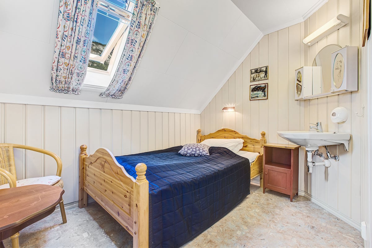 A bedroom with a wooden bed, a bedside table, a chair, a window with patterned curtains, a wall-mounted mirror, and a small wooden cabinet.