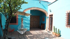 Un espace extérieur aménagé en patio, avec un mur bleu, une porte en bois et une arche en briques. On y trouve deux chaises en métal et un pot de fleurs contenant des cactus.