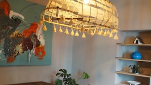 Un espace repas comprenant une table en bois, des chaises blanches, un luminaire suspendu, un tableau mural et des étagères ornées d’objets décoratifs.