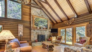 A log cabin interior with a stone fireplace, leather furniture, and large windows offering views of the forest. - Black Bear Lodge by Vtrips (Cosby)