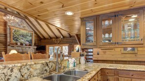 A rustic kitchen with wooden cabinets, granite countertops, and a stone fireplace. - Black Bear Lodge by Vtrips (Cosby)