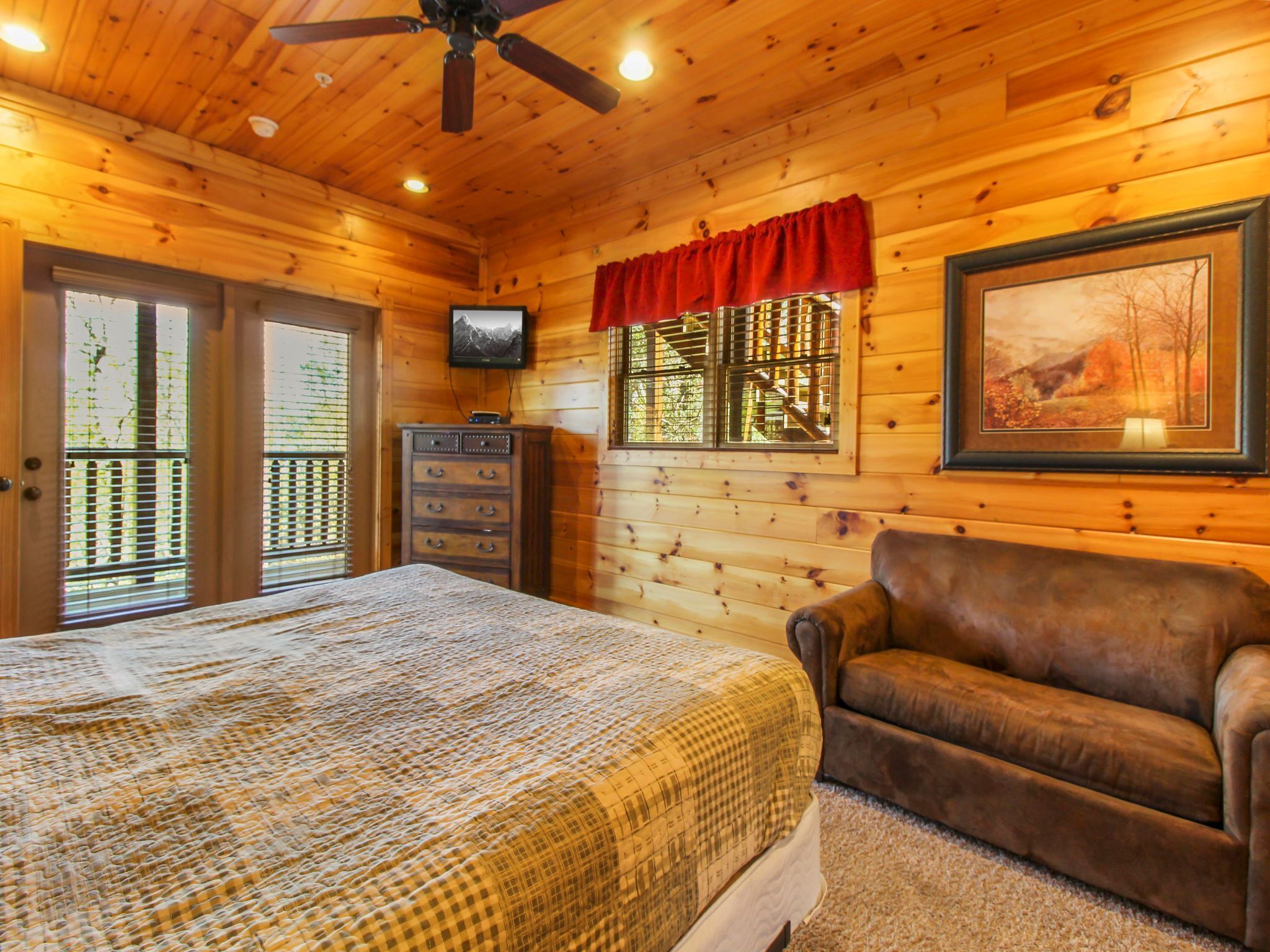A wooden cabin bedroom with a bed, a television, a dresser, a window with a red valance, and a framed picture on the wall.