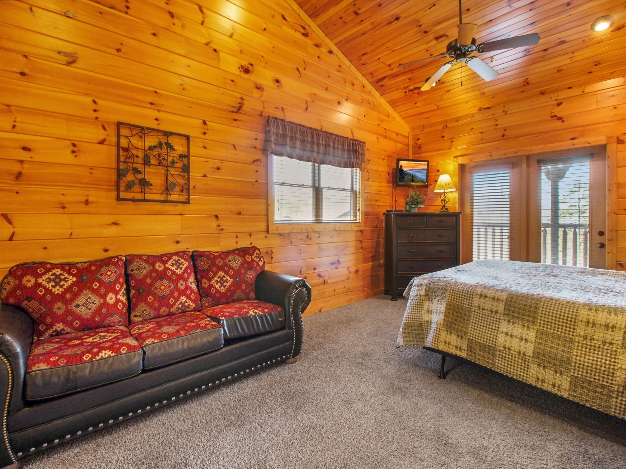 A cozy cabin room with a wooden ceiling, a leather sofa, a bed, a dresser, and a window with blinds.