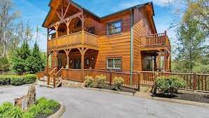 A two-story log cabin with a balcony on the second floor, surrounded by a stone retaining wall and a landscaped garden. - Big Sky Lodge II by Jackson Mountain Rentals (Gatlinburg)