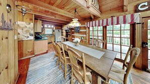 A wooden cabin interior with a dining table, chairs, a kitchen area, and large windows. - Bear Cave Haus by Jackson Mountain Rentals (Gatlinburg)