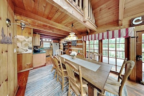 A wooden cabin interior with a dining table, chairs, a kitchen area, and large windows. - Bear Cave Haus by Jackson Mountain Rentals (Gatlinburg)