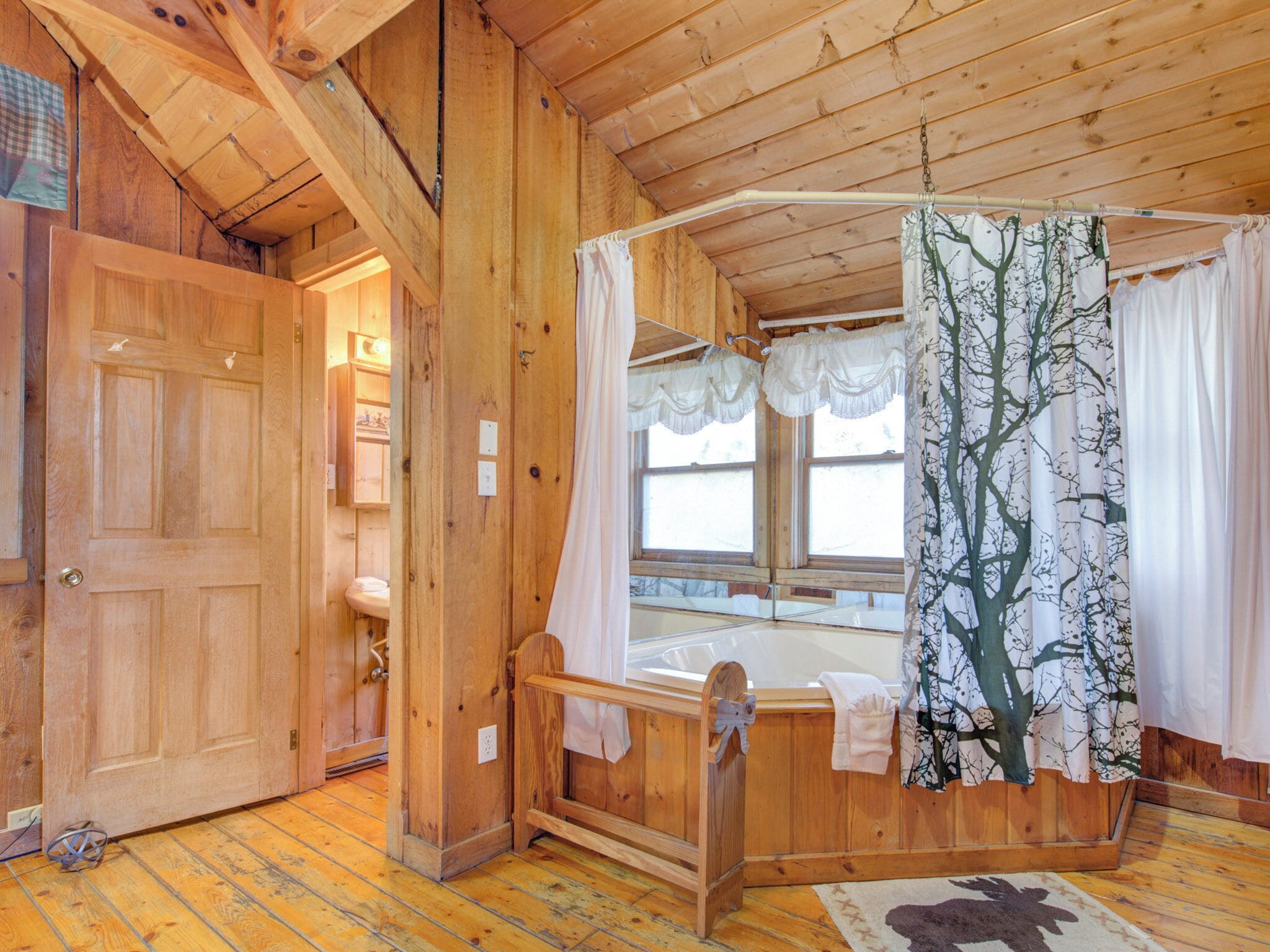 A wooden cabin interior with a wooden door, a bathroom with a sink, a wooden bed with a canopy, and a window with a tree-patterned curtain.