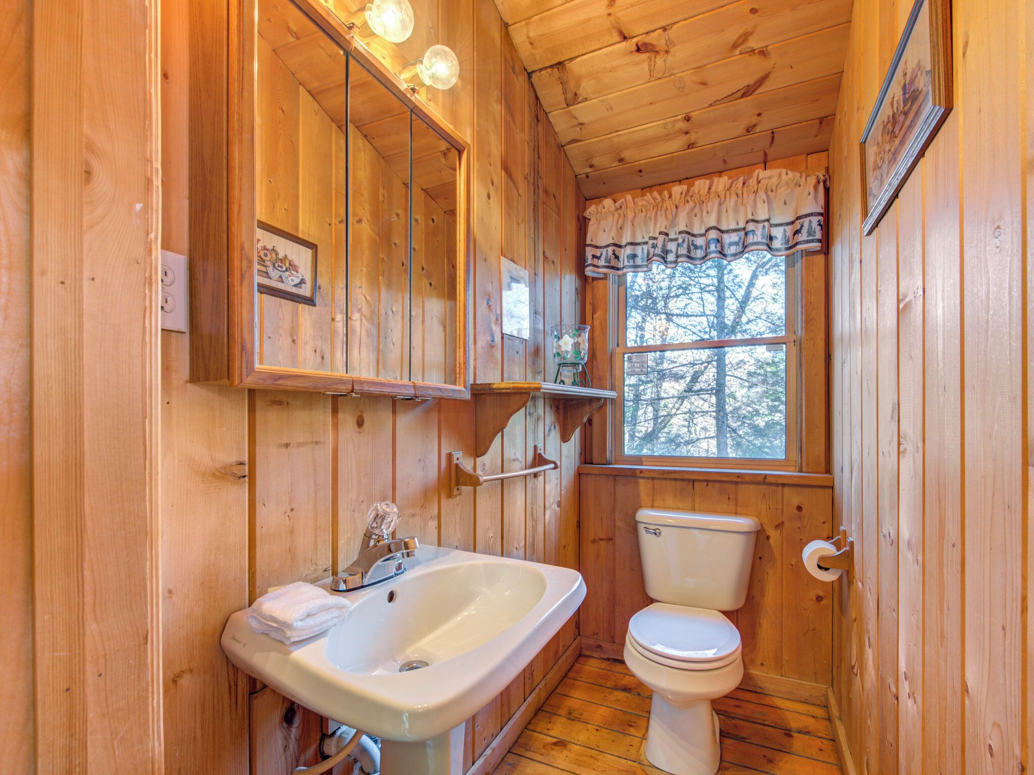 Une salle de bain rustique, aménagée dans une cabane en bois, avec un lavabo, des toilettes et une fenêtre donnant sur la forêt.