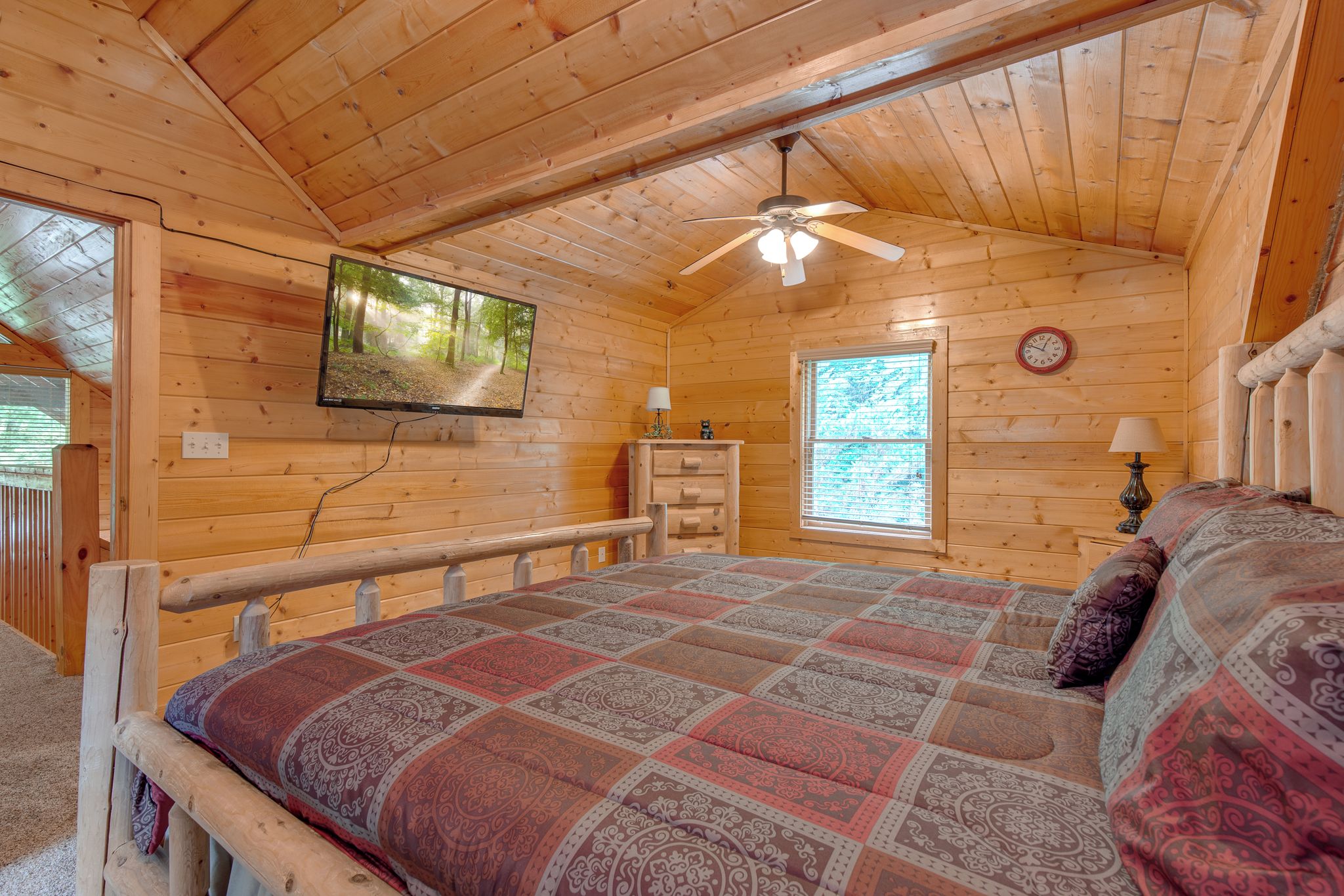A cozy wooden cabin room with a bed, a ceiling fan, a TV mounted on the wall, a window, and a dresser.