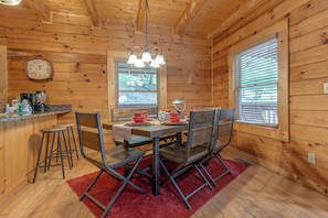 A wooden cabin interior with a dining table set for four, a kitchen area with a clock, and a window with blinds.