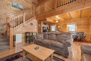 A wooden cabin interior with a staircase, a living room with a sofa and coffee table, a kitchen area, and a dining table with chairs.