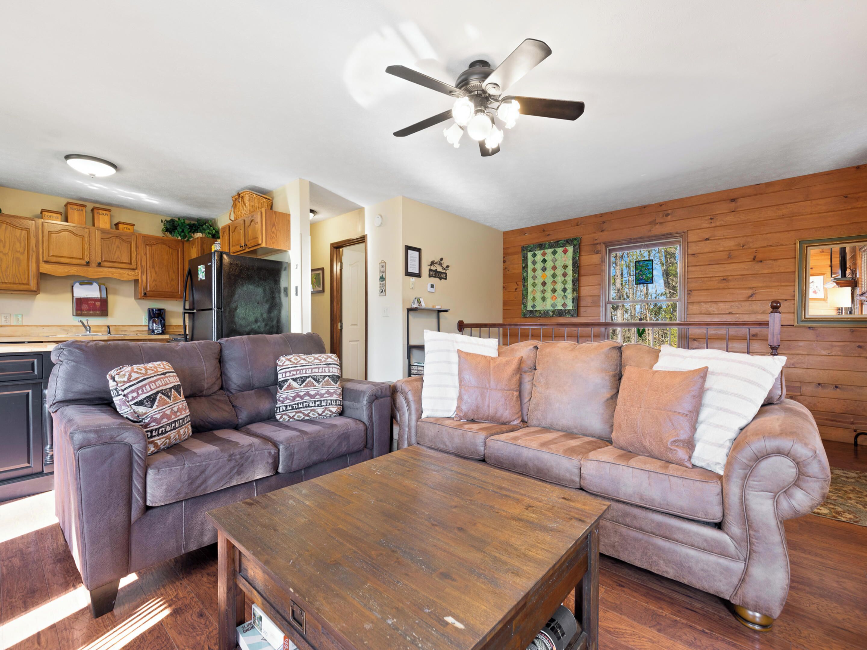 A living room with two sofas, a coffee table, wooden cabinets, a ceiling fan, and a window with a view of trees.