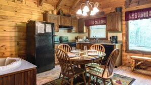 Une cuisine rustique aménagée dans une cabane en bois, avec une table à manger, des chaises, un réfrigérateur et une fenêtre équipée de stores.