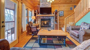 A cozy cabin interior with a stone fireplace, wooden walls, a ceiling fan, and a large window overlooking a deck. - Dew South by Jackson Mountain Rentals (Gatlinburg)
