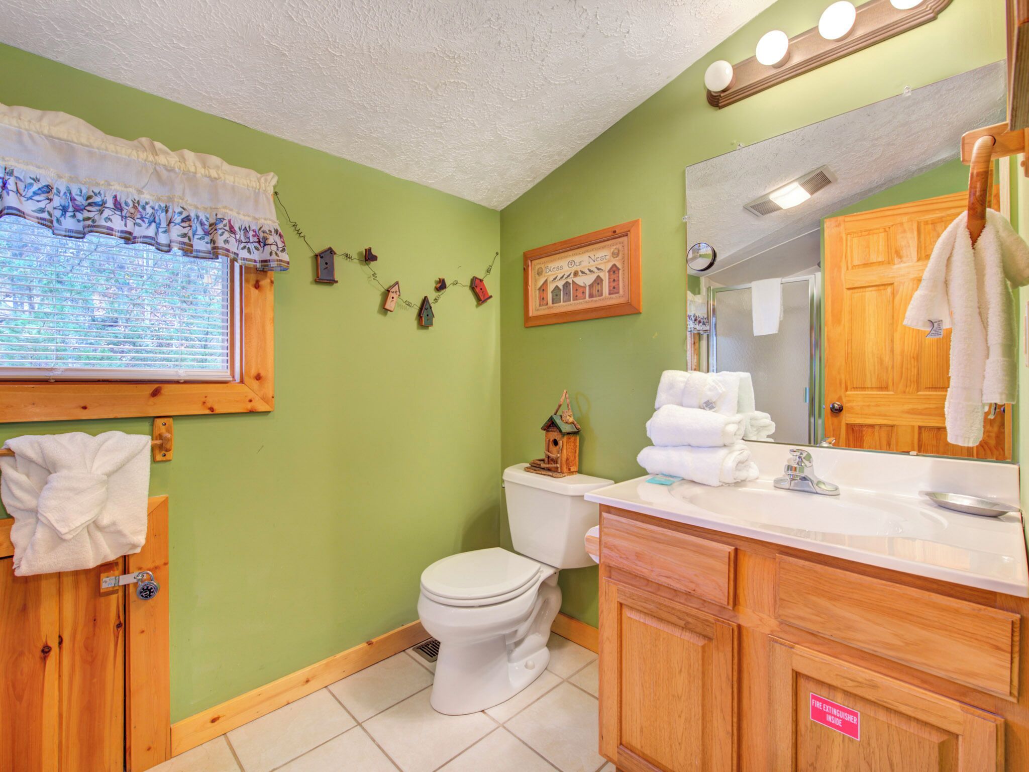A bathroom with a toilet, sink, and mirror. There is a window with blinds, a towel rack, and a small birdhouse decoration on the wall.