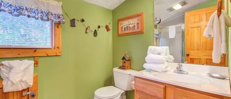 A bathroom with a toilet, sink, and mirror. There is a window with blinds, a towel rack, and a small birdhouse decoration on the wall.
