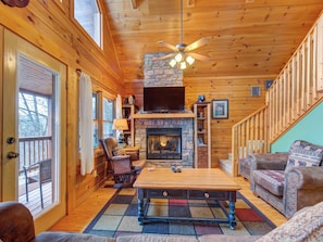 A cozy cabin interior with a stone fireplace, wooden walls, a ceiling fan, and a large window overlooking a deck.