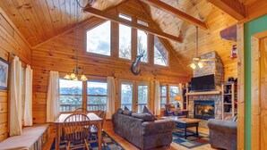 A cozy cabin interior with a dining area, sofa, fireplace, and large windows offering mountain views. - Dew South by Jackson Mountain Rentals (Gatlinburg)