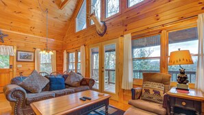 A cozy cabin interior with a wooden ceiling, large windows, a sofa, a coffee table, and a fireplace. - Dew South by Jackson Mountain Rentals (Gatlinburg)