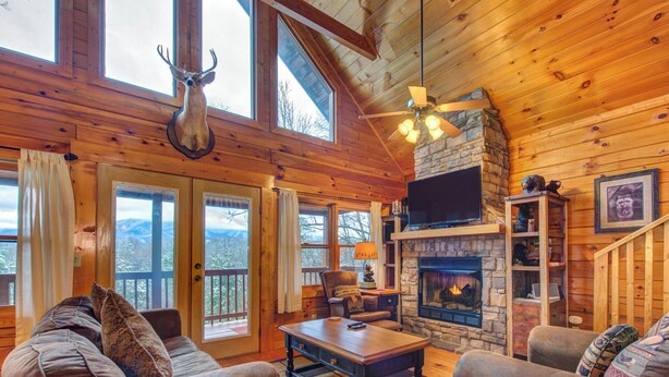 A cozy cabin interior with a stone fireplace, wooden furniture, and a mounted deer head. - Dew South by Jackson Mountain Rentals (Gatlinburg)