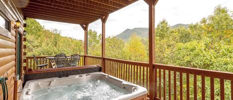 A wooden deck with a hot tub, outdoor seating, and a view of a forested mountain range.