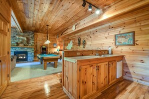 A wooden cabin interior with a pool table, a fireplace, and a kitchen area with wooden cabinets. - A Majestic View III by Jackson Mountain Rentals (Townsend)