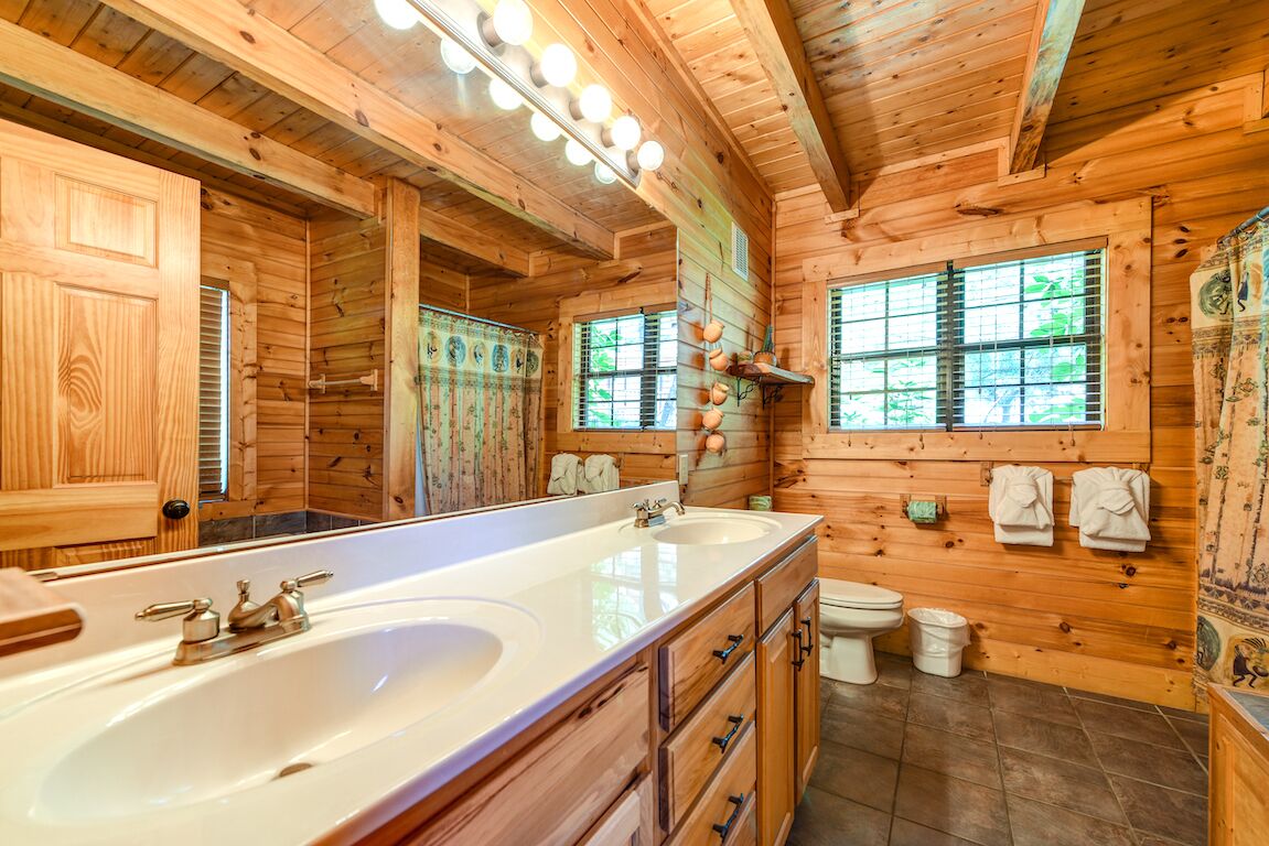 A wooden cabin bathroom with a large mirror, two sinks, a toilet, and a window.
