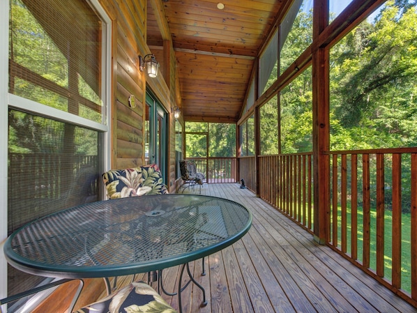 A covered porch with a round table and chairs, a wooden deck, and a view of the surrounding forest.
