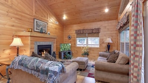 Un acogedor interior de cabaña de madera con chimenea, dos sofás, un televisor y una mesa de comedor.