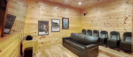 Une pièce aux murs et au plafond en bois, avec un canapé en cuir noir et plusieurs chaises noires. Des affiches encadrées ornent les murs.